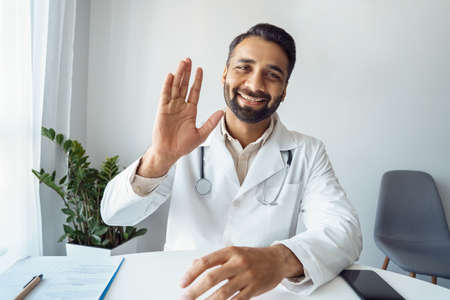 Headshot portrait of positive smiling indian man doctor in uniform saying hiの写真素材