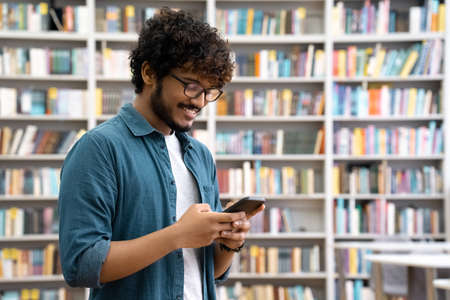 Side view portrait of happy young Indian student chatting by phone in libraryの写真素材