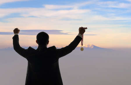 Businessman holding golden medal while staring peak of mountain. Concept of success of startup business and revenue, income, profit celebration.の写真素材
