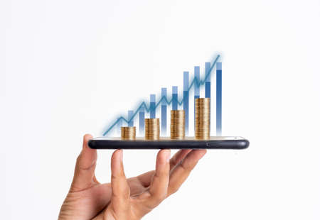 Close up photo of businessman holding smartphone with coin stacks and graphics as a symbol of trading money digitally.の写真素材