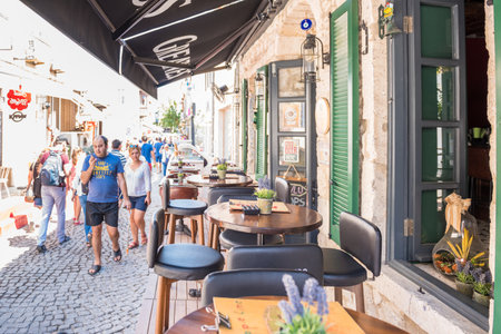 Unidentified People walk at a street with cafe and restaurant with tables and chairs  around at Alacati Town,a popular destination for traveling and vacation in Izmir,Turkey.26 August 2017.のeditorial素材