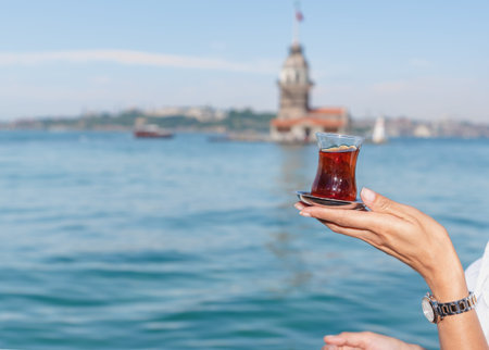 Beautiful woman traveler holds traditional Turkish tea glass in background of Maiden Tower,a popular...