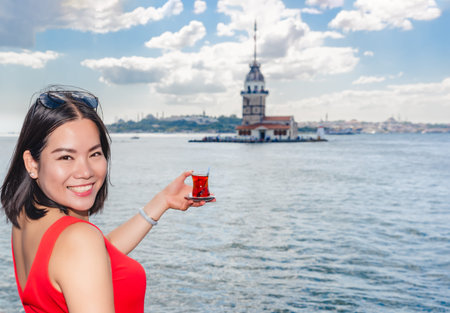 Beautiful Chinese woman holds traditional Turkish tea with Maiden's Tower view on background in Istanbul,Turkeyの写真素材