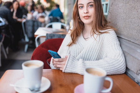 Beautiful attractive young girl in jumper holds and uses smart phone and texting while sitting a cafe.の写真素材