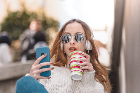 Portrait of beautiful attractive young trendy girl with headphones and sunglasses uses smart phone...