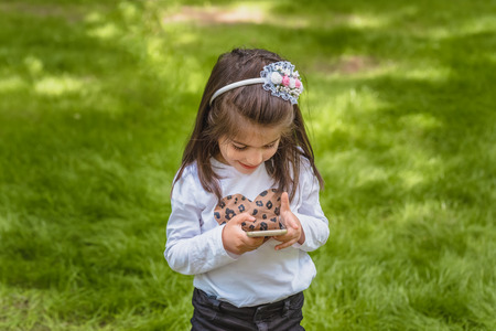 Adorable four years old cute little girl in casual clothes holds and texts mobile phone while smiling at outdoor in parkの写真素材