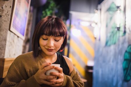 Beautiful young sexy girl sits and drinks coffee alone at a trendy cafe or bar at night.の写真素材