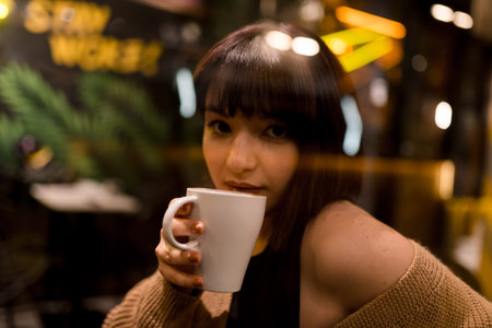 Beautiful young girl sits and drinks coffee at a trendy cafe or bar at night with window...
