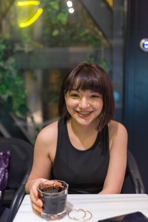 View of beautiful young girl in black dress while sitting at a table with a glass of beverage in cafe or bar.の写真素材
