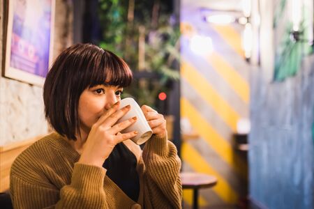 Beautiful young sexy girl sits and drinks coffee alone at a trendy cafe or bar at night.の写真素材