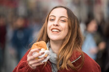 Beautiful young cute girl in fashionable clothes enjoys fast food hamburger whilewalking at a crowded streetの写真素材