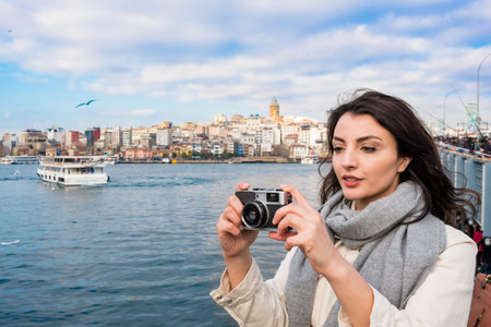 Beautiful woman in fashionable modern clothes takes photos with retro vintage camera with view of Galata Tower,Galata Bridge and Bosphorus in Istanbul,Turkeyの写真素材
