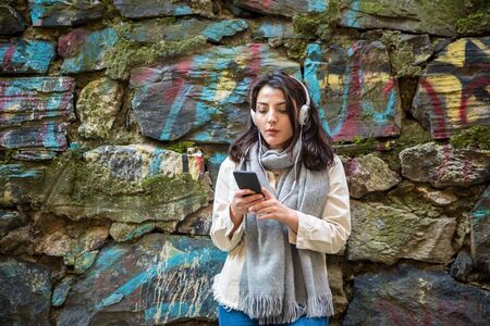Beautiful young girl in fashionable clothes listens music and uses smart phone with stone background while standingの写真素材