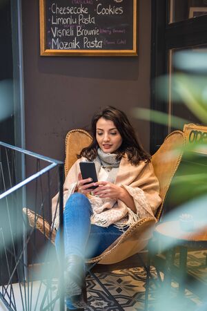 Beautiful young girl in fashionable clothes reads or texts at smartphone while sitting at a cafe outside.Woman Lifestyle conceptの写真素材