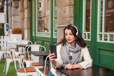 Beautiful young girl in fashionable clothes listen music with headphones and takes selfie while sitting at a cafe outside.Lifestyle conceptの写真素材