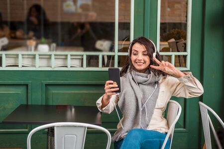 Beautiful young girl in fashionable clothes listen music with headphones and takes selfie while sitting at a cafe outside.Lifestyle conceptの写真素材