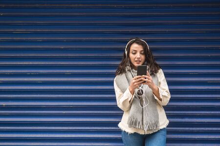 Beautiful young girl in fashionable clothes listens music and uses smart phone with blue background while standingの写真素材