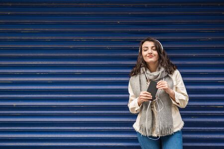 Beautiful young girl in fashionable clothes listens music and uses smart phone with blue background while standingの写真素材