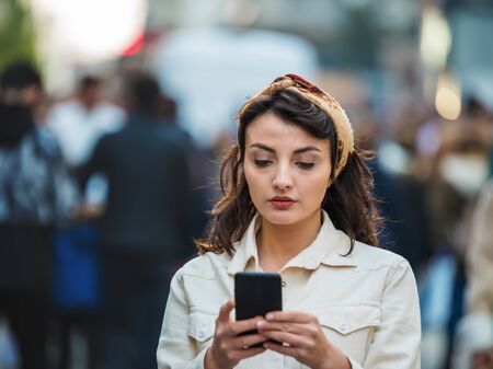 Beautiful young girl in fashionable clothes uses smartphone  while walking in crowded people at busy street.Girl on the street among the crowd uses her phone.の写真素材