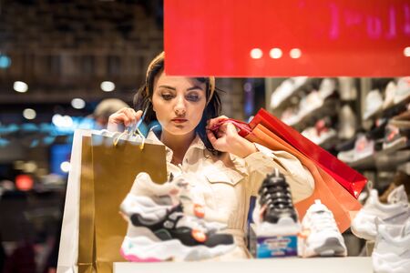 Beautiful woman in fashionable clothes looks different kind of sport shoes while shopping in store with bags.Shopping, lifestyle conceptの写真素材