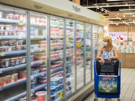 Young woman with shopping cart wears medical mask looks at produces in dairy shopping mall during the Coronavirus Covid-19 epidemic 2020の写真素材