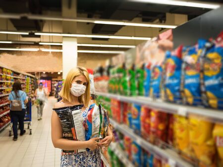 Young beautiful woman with medical mask holds potato chips packs while shopping and stockpiling in supermarket. Shopping concept during pandemic corona,covid19.の写真素材