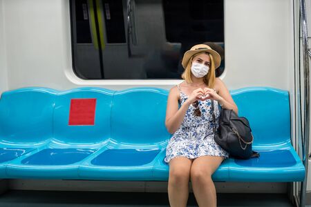 Portrait of happy beautiful woman wearing protective face mask and keeps social distance by sitting alone with love heart gesture and smile during covid-19 or coronavirus outbreak .の写真素材