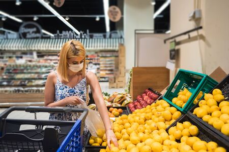 Young woman with shopping cart wears medical mask and buys lemons and fruits. Shopping during the Coronavirus Covid-19 epidemic 2020の写真素材