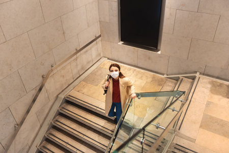 Beautiful girl wearing protective disposable mask and fashionable clothes walking down at stairs....