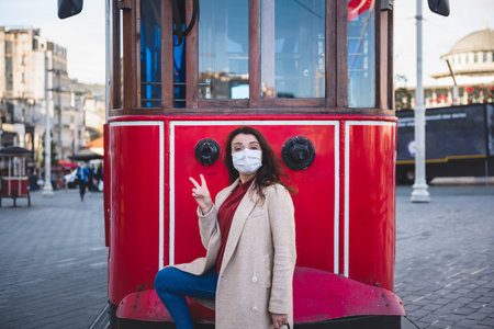 Beautiful girl wearing protective medical mask and fashionable clothes poses with red tram at istiklal street in Istanbul,Turkey. New normal lifestyle concept.の写真素材