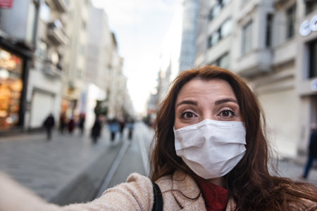 Beautiful girl wearing protective medical mask and fashionable clothes takes selfie with a smart phone at street. New normal lifestyle concept.の写真素材