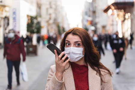 Beautiful girl wearing protective medical mask and fashionable clothes speaks with a smart phone at street. New normal lifestyle concept.の写真素材