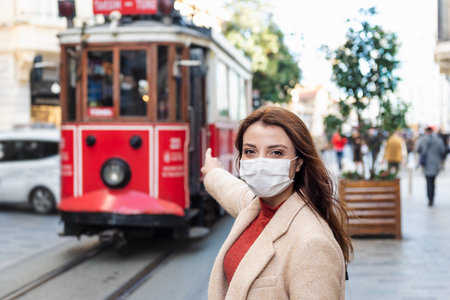 Beautiful girl wearing protective medical mask and fashionable clothes shows nostalgic tramway at istiklal street, Istanbul,Turkey. New normal lifestyle concept.の写真素材