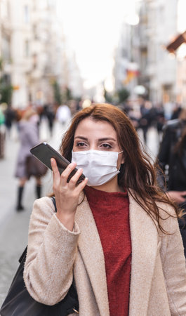 Beautiful girl wearing protective medical mask and fashionable clothes speaks with a smart phone at street. New normal lifestyle concept.の写真素材