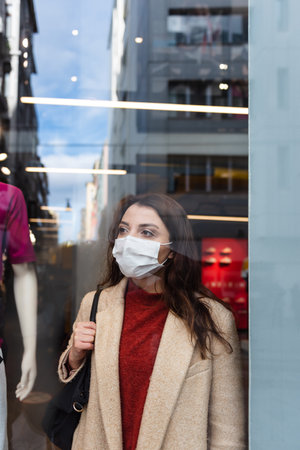 Beautiful girl wearing protective medical mask and fashionable clothes looks at behind of showcase. New normal lifestyle concept.の写真素材