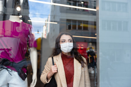 Beautiful girl wearing protective medical mask and fashionable clothes looks at behind of showcase. New normal lifestyle concept.の写真素材