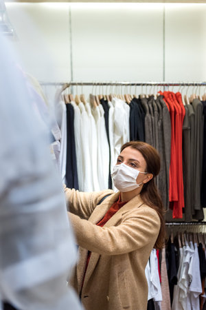 Beautiful girl wearing protective medical mask and fashionable clothes looks at clothes at store. New normal lifestyle concept.の写真素材