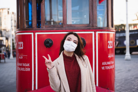 Beautiful girl wearing protective medical mask and fashionable clothes poses with red tram at istiklal street in Istanbul,Turkey. New normal lifestyle concept.の写真素材