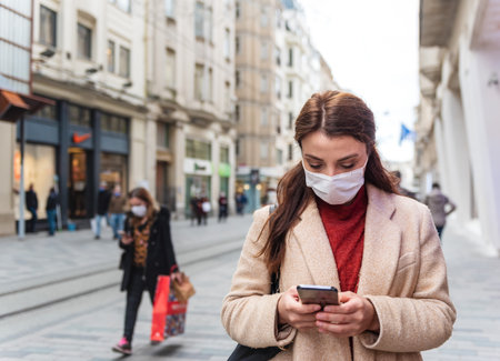Beautiful girl wearing protective medical mask and fashionable clothes uses smart phone at street. New normal lifestyle concept.の写真素材