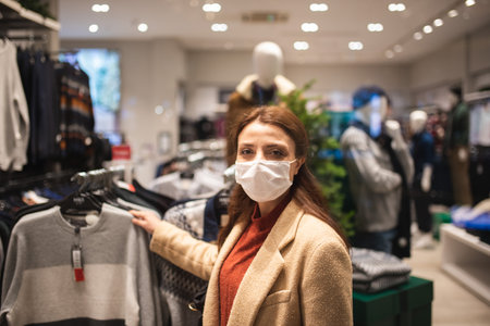 Beautiful girl wearing protective medical mask and fashionable clothes looks at behind of showcase. New normal lifestyle concept.の写真素材