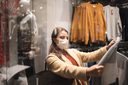 Beautiful girl wearing protective medical mask and fashionable clothes looks at behind of showcase. New normal lifestyle concept.の写真素材