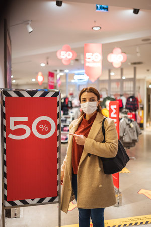 Beautiful girl wearing protective medical mask and fashionable clothes shows sale,discount clothes store. New normal lifestyle concept.の写真素材
