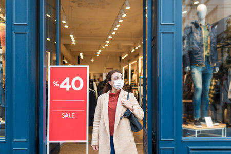 Beautiful girl wearing protective medical mask and fashionable clothes shows sale,discount clothes store. New normal lifestyle concept.の写真素材