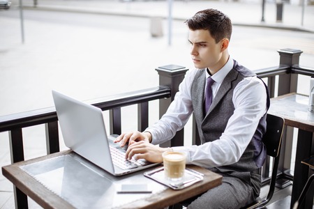 Handsome businessman wearing suit and using modern laptop outdoors, successful manager working in cafe during break and searching information in internet on his notebook computerの写真素材