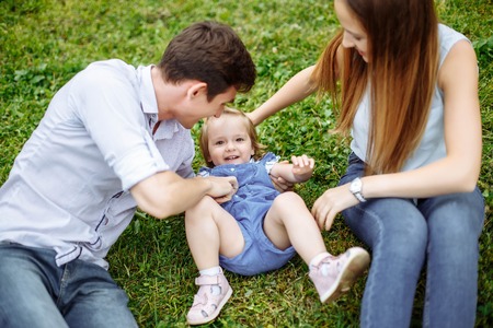 Portrait happy family of three. Young parents play with their little daughter sitting on the lawn in the Park.の写真素材