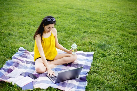 Beautiful young woman sitting on the green grass in park with her laptop. Student girl looking at the computerの写真素材