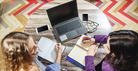 Two young women doing records in a notebook in an office top view. Laptop, glasses, smart phone on the table.の写真素材