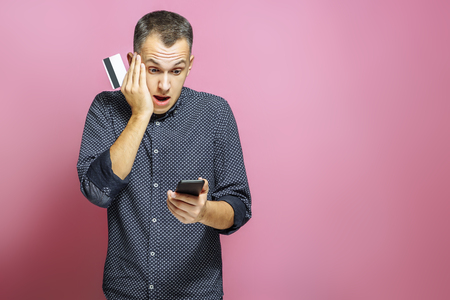 Scared man holding credit card and smartphone on pink background.の写真素材