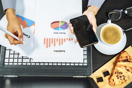 Female hands holding a smartphone and making notes in graphs on paper. Business breakfast concept.