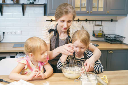 Happy family young mother and two little daughters preparing dough for pancakes or cookies together in the kitchen.の写真素材